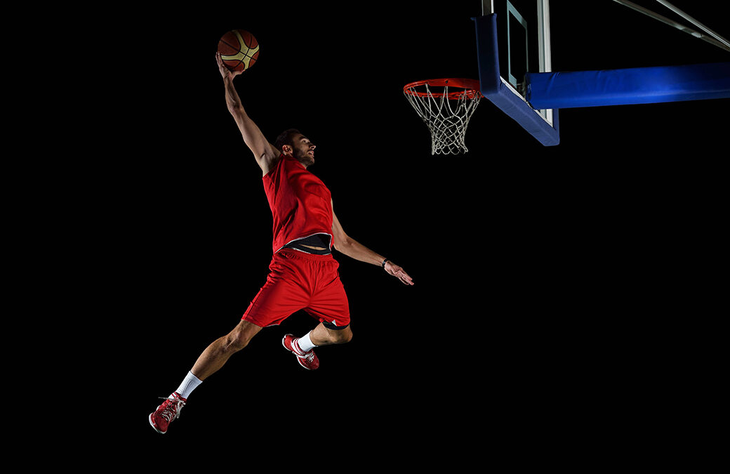 A man in a red basketball uniform in mid-air dunking a basketball against a darkened stadium.