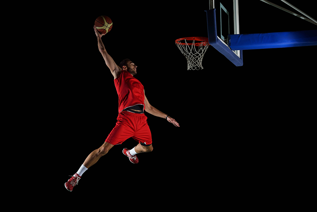 A man in a red basketball uniform in mid-air dunking a basketball against a darkened stadium.