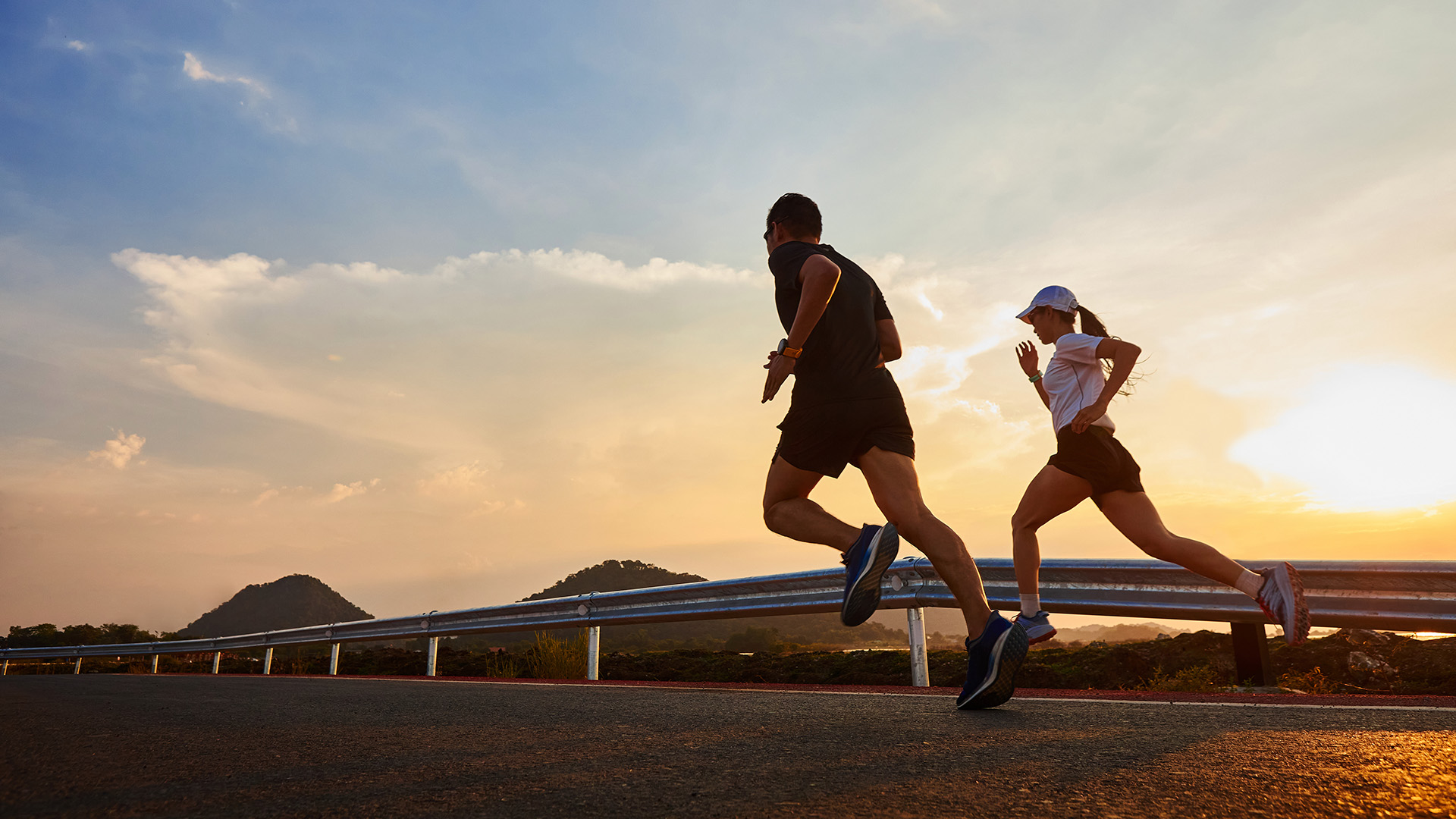 A man and a woman running on a road next to each other against a sunset backdrop.