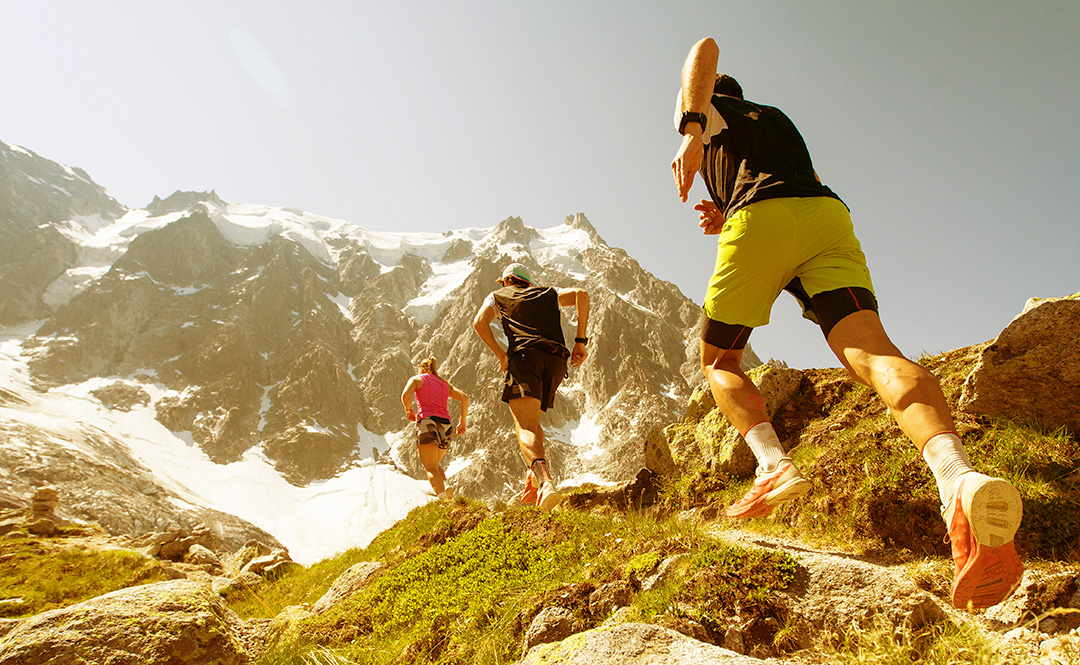 Three trail runners on an open mountain trail heading towards a range of snowy peaks