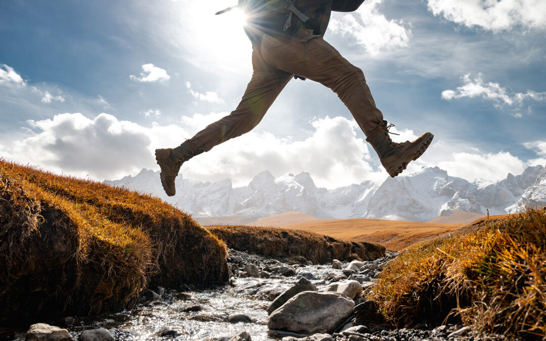 A male hiker leaping over a flowing brook lined by red grass and silhouetted against a blue sky