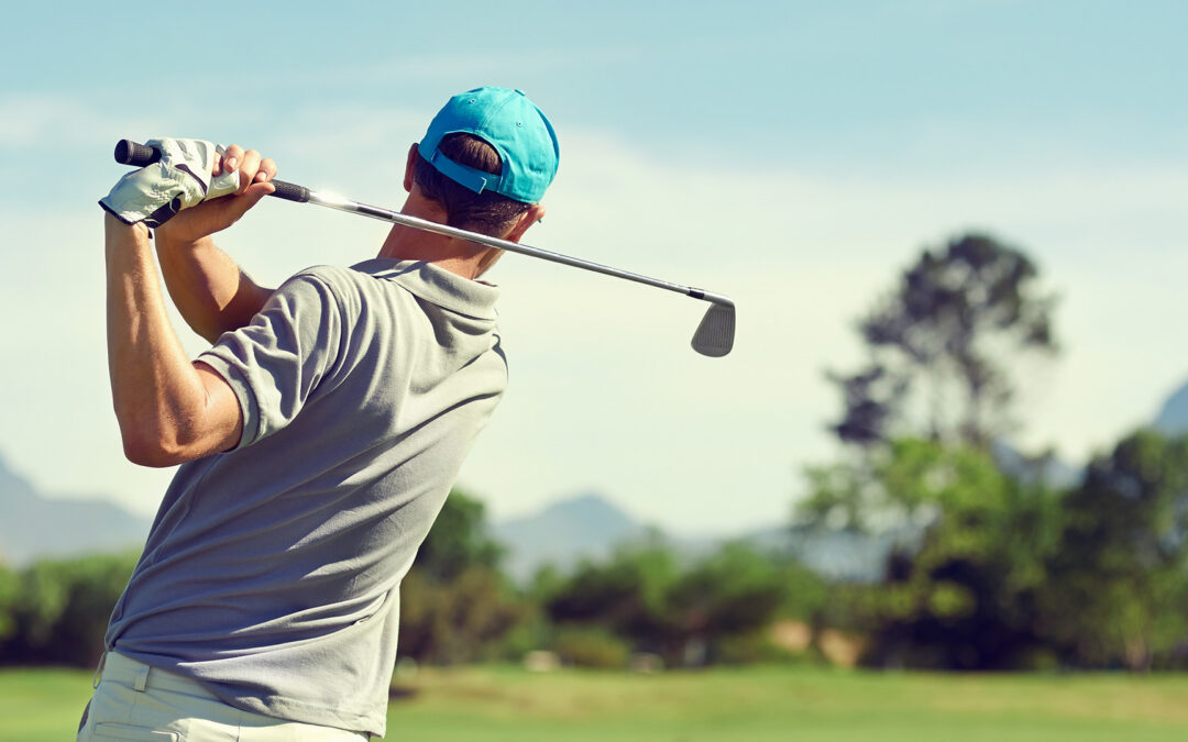 A back view of a golfer taking a large swing on a course with a line of trees and mountains in the background.