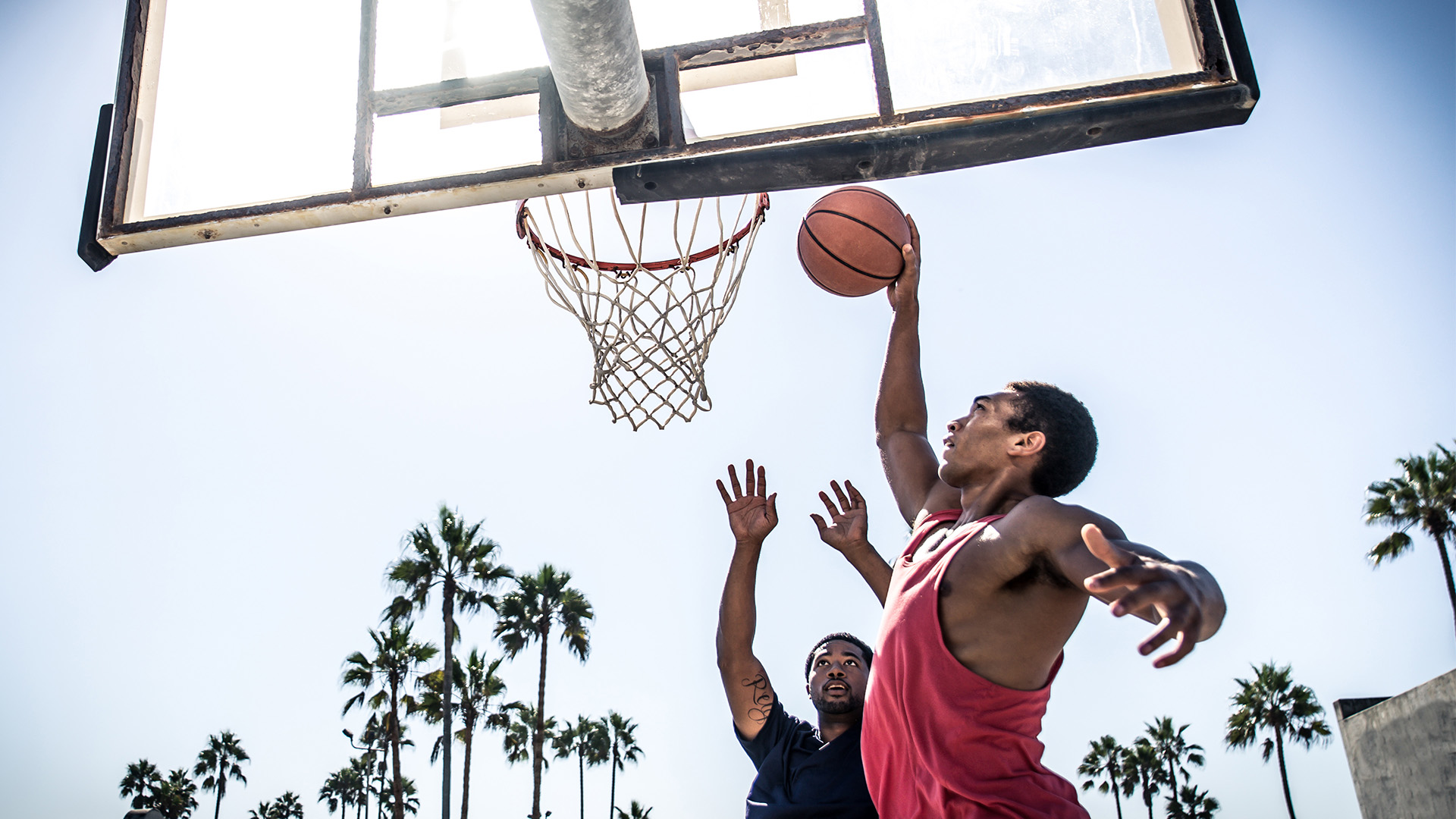 Two amateur basketball players on an outdoor court with palm trees in the background. One is reaching up to dunk the ball while the other is trying to block.